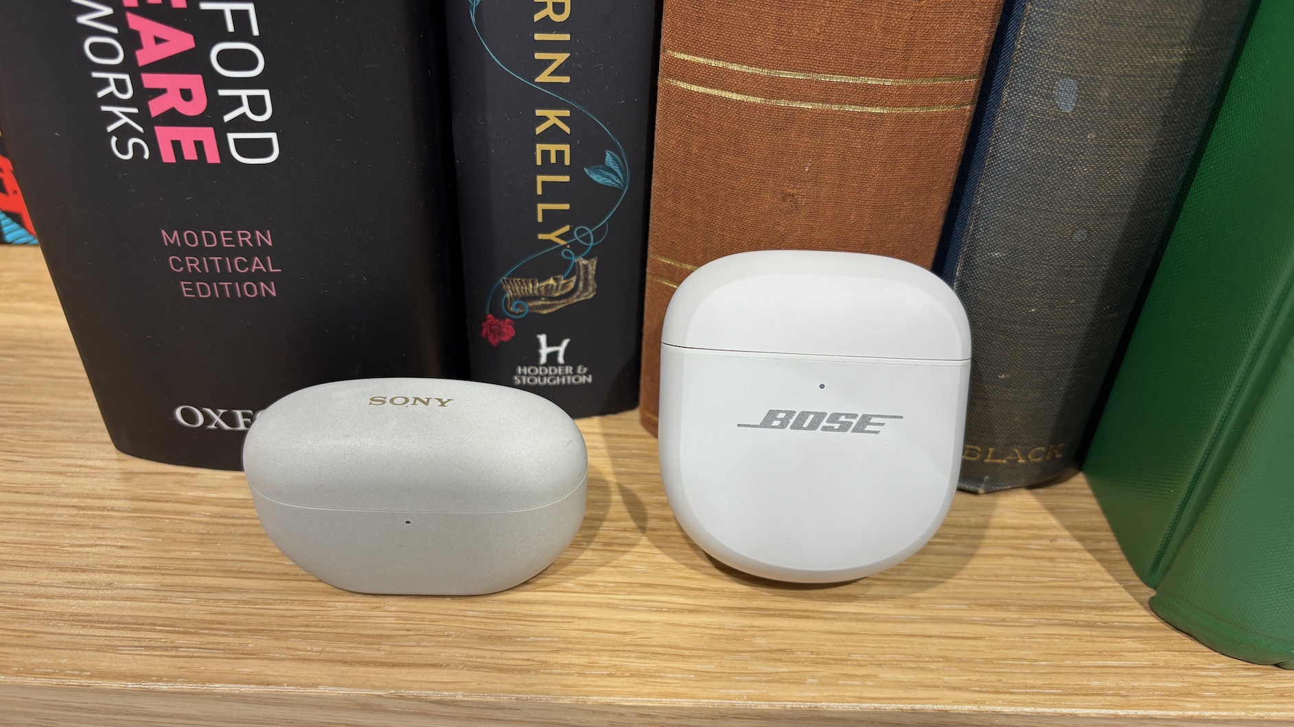The Bose QuietComfort Ultra Earbuds (2nd Gen) and Sony WF-1000XM5 in their charging cases on a shelf in front of a row of books.