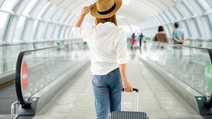 A woman in a straw hat walks through an airport toting a suitcase.