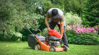 man in garden pouring petrol from red container in orange and black petrol lawnmower on lawn