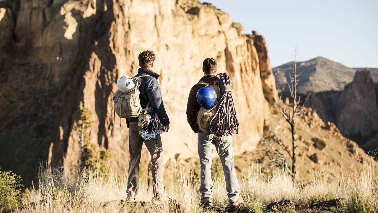 Two men with climbing equipment consider scaling a cliff.