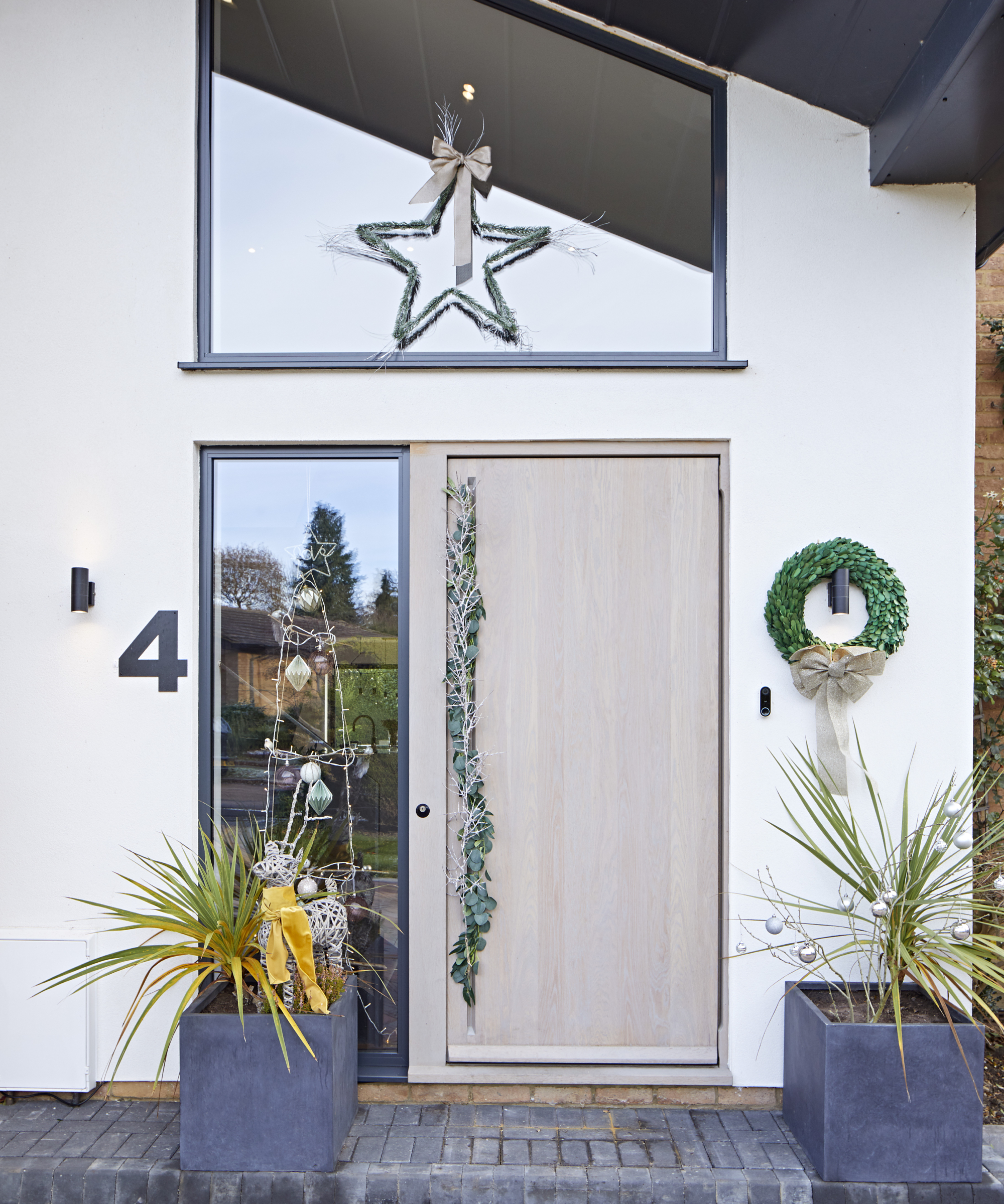 light wooden front door with star on glass panel above, wreath hanging over outdoor light and handle decorated with a garland