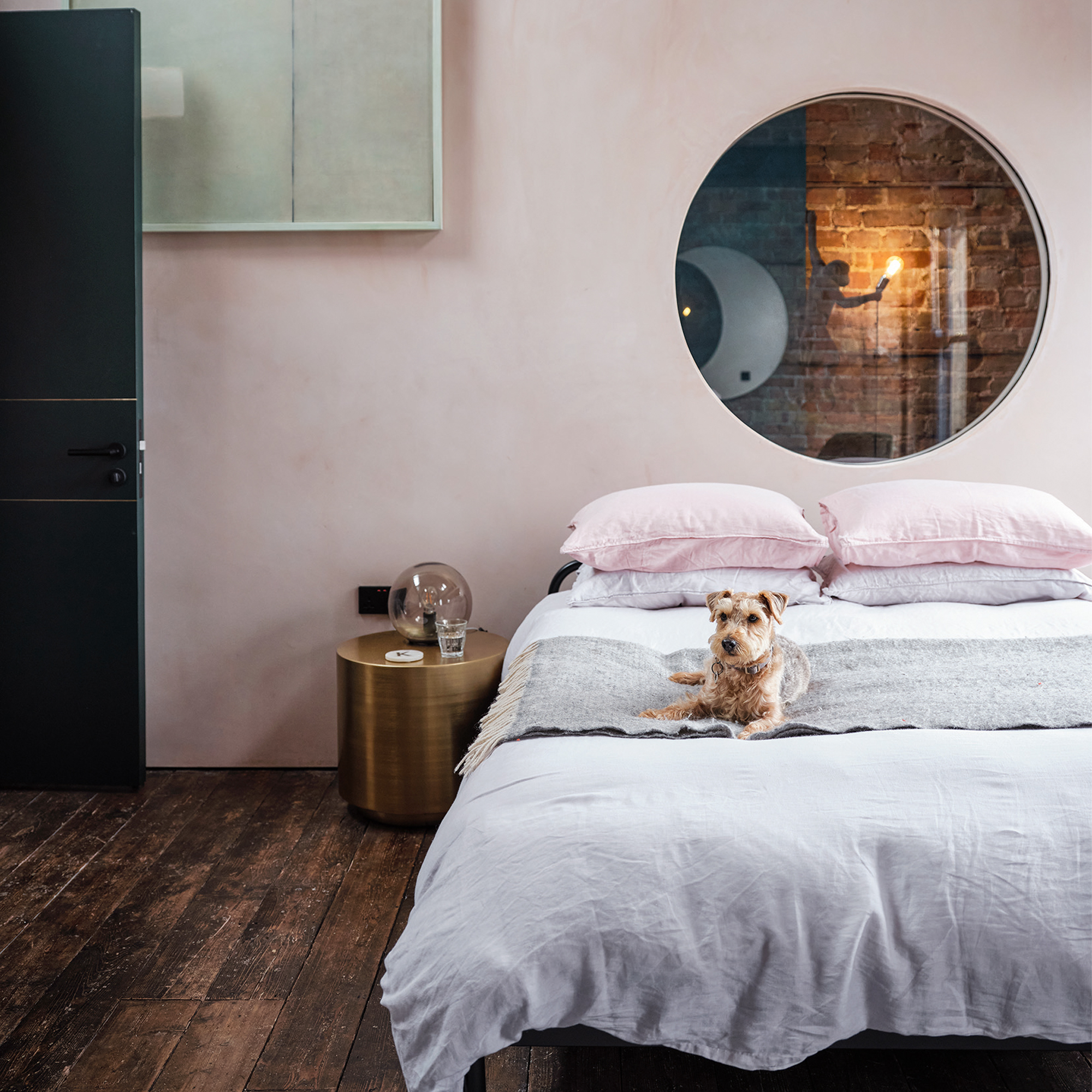 A pale pink bedroom with round mirror hanging over the bed and a small dog lying on the bed
