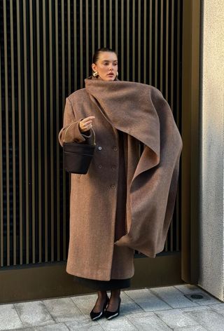 The winter capsule wardrobe item, an extra-long scarf, is shown in a photo of a woman standing outside wearing gold drop earrings with a long brown coat styled with a long scarf, black skirt, sheer tights, black kitten pumps, and black bucket bag