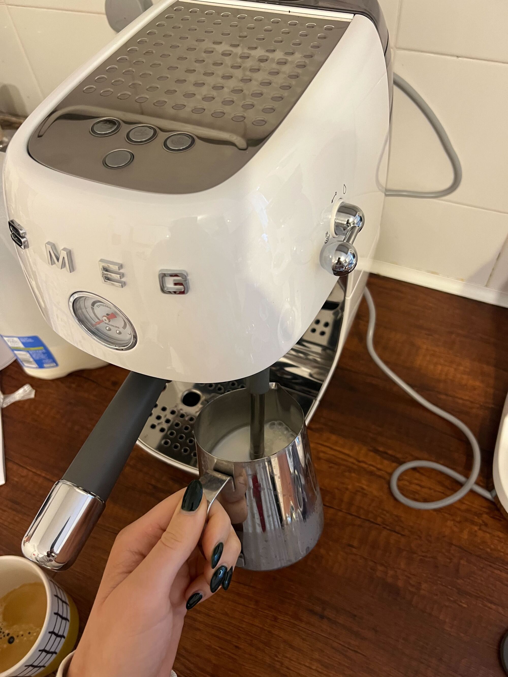 Image of a white Smeg cold brew machine and a hand steaming milk in a stainless steel milk jug on a wooden countertop with white tile backsplash