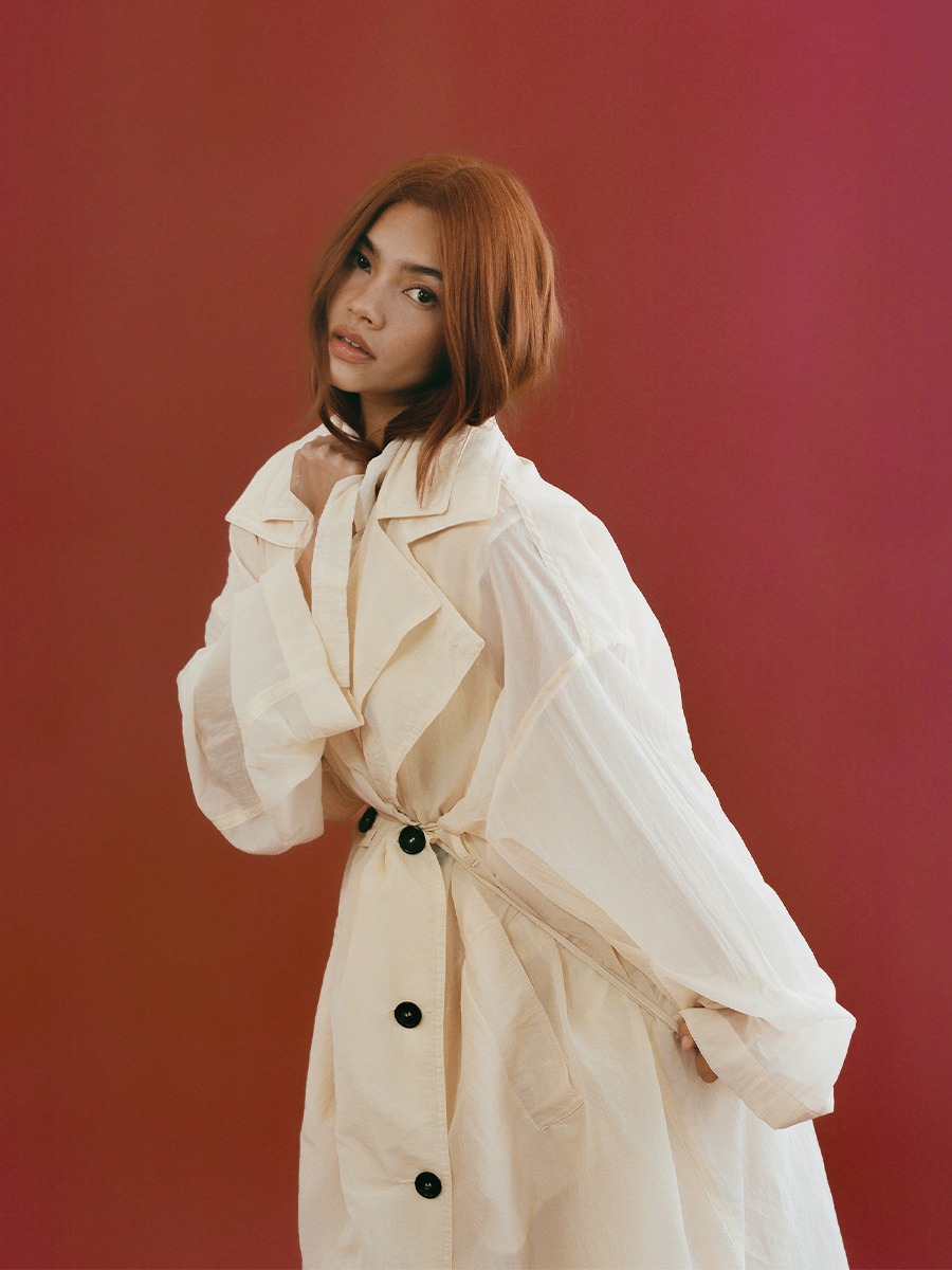 Myra Molloy photographed by David Dickenson. She is in front of a red backdrop wearing an oversize white trench coat. Her hair is tucked into the coat and she is tightening the waist strap.