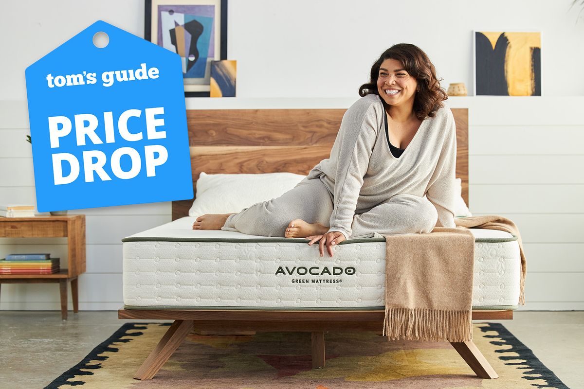 A woman sits on the end of an Avocado Green Mattress in a bedroom, a Tom&#039;s Guide price drop deals graphic (left)