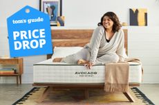 A woman sits on the end of an Avocado Green Mattress in a bedroom, a Tom's Guide price drop deals graphic (left)