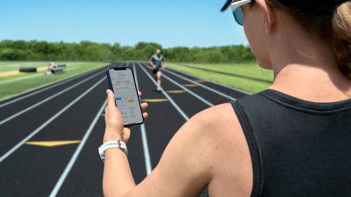 A coach standing on a running track looks at the Garmin Clipboard app on a smartphone, a runner in the background is running towards them