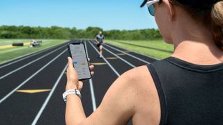 A coach standing on a running track looks at the Garmin Clipboard app on a smartphone, a runner in the background is running towards them