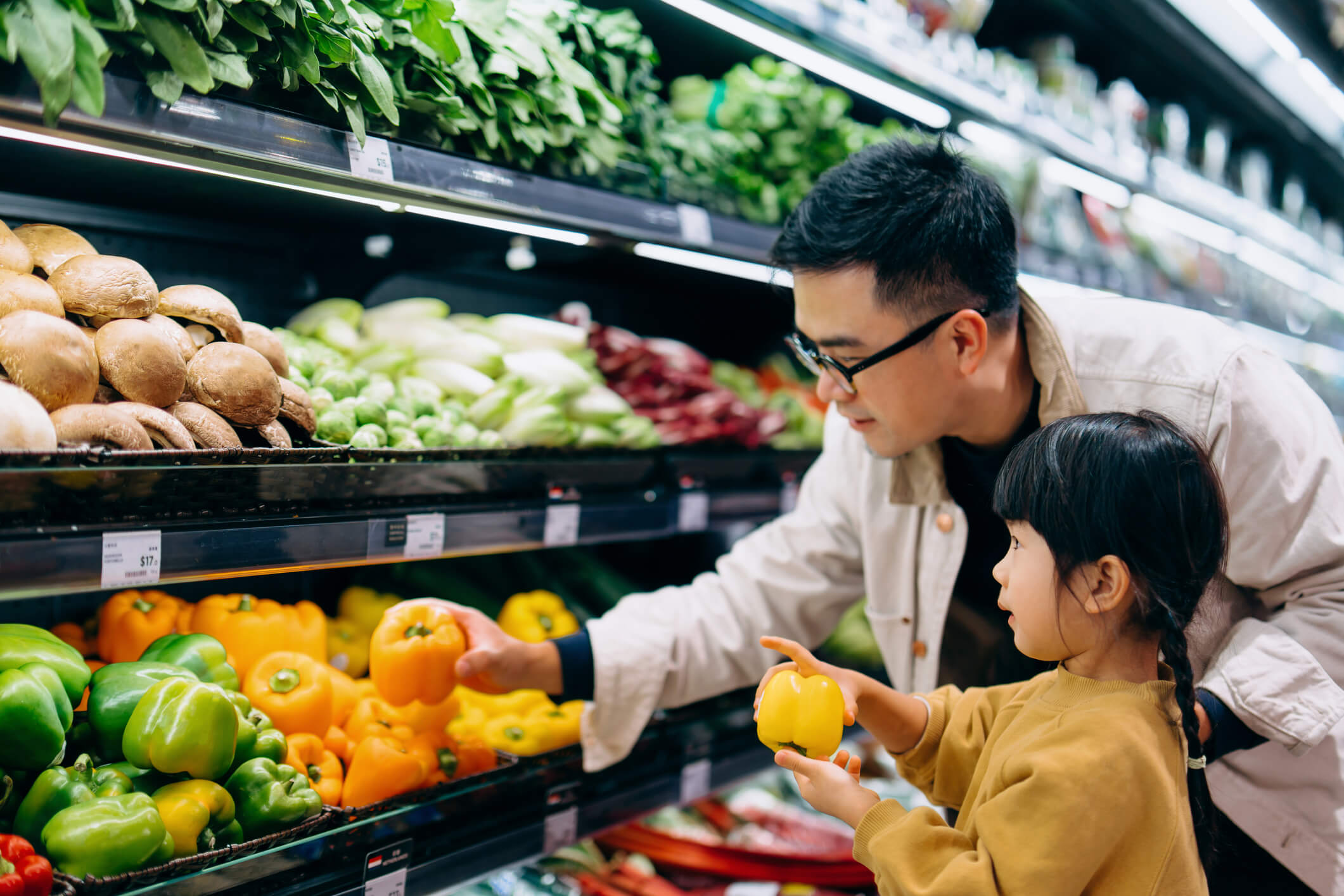 Young Asian father and her little daughter grocery shopping in supermarket. They are choosing fresh organic bell peppers together along the produce aisle.