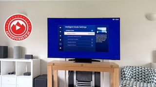 The Samsung S95C photographed on a wooden table in a living room. On the screen is the Intelligent Mode Settings menu, and in the corner of the photo is a badge that says &#039;Adventures in AV&#039;.