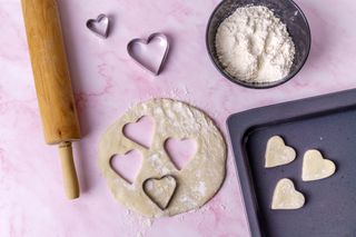 A pink surface with a rolling pin, a bowl of flour, and heart cut-out biscuits.