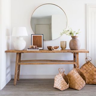 a white living room with a rustic feel, a wooden console table holding a white lamp and other items, with a large circle gold rimmed mirror above, with three brown weaved baskets on the brown floor