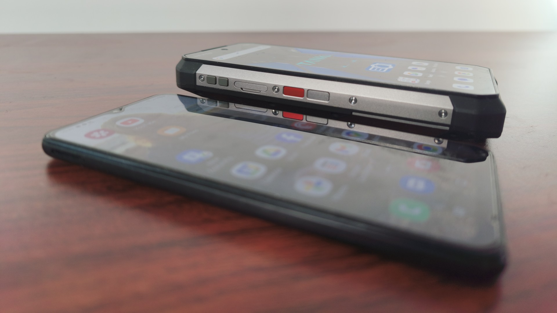 A thick, rugged smartphone with visible side buttons is resting next to a thinner, standard smartphone on a wooden surface, showing the size difference.
