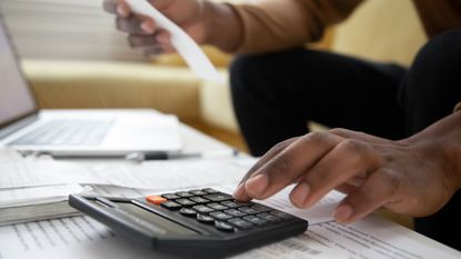 A man's fingers tap on a calculator with financial paperwork showing in the background. 