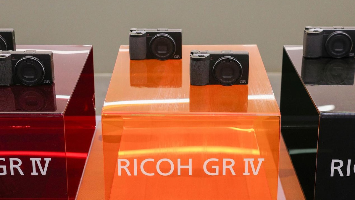 Three Ricoh GR IV cameras are displayed on colorful pedestals. The left is red, the middle orange, and the right black, each labeled &quot;Ricoh GR IV&quot;