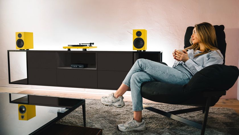 A woman relaxing in an armchair clasping a mug, gazing admiringly at a yellow turntable and speakers on a hi-fi cabinet.