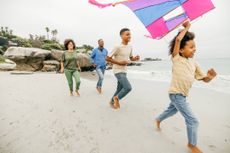 A family are running on the beach. The child in the lead is holding the string to a colourful kite.