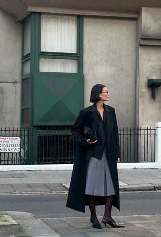 A cute work winter outfit is shown in a photo of a woman standing on the sidewalk in London wearing a long black wool coat over a black button-down shirt styled with a gray pleated midi skirt, black tights, black pointed slingbacks, and a black clutch bag