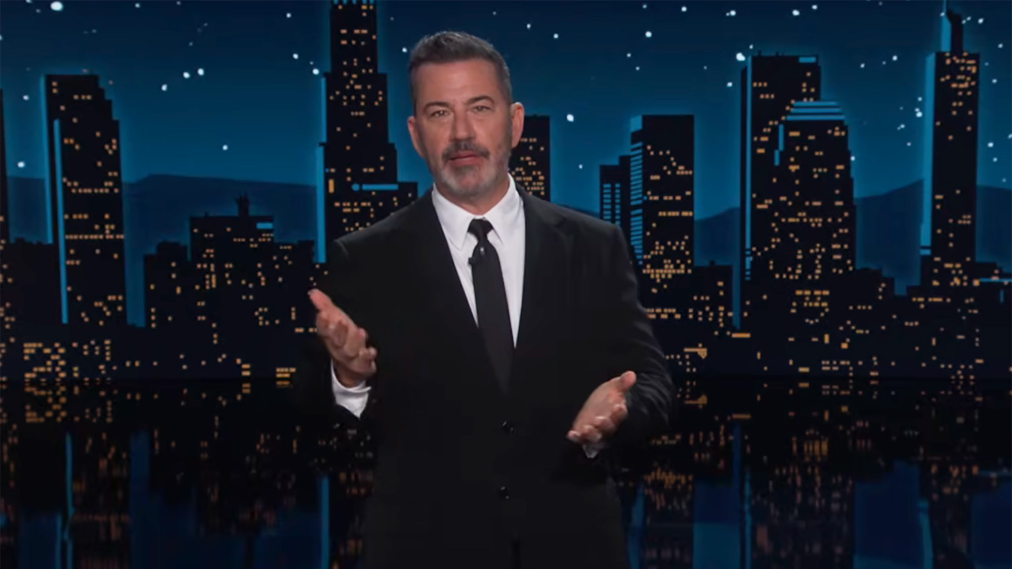 Jimmy Kimmel holds up his hands while talking about summer events on Jimmy Kimmel Live. The backdrop is the city and he's wearing a suit and tie.