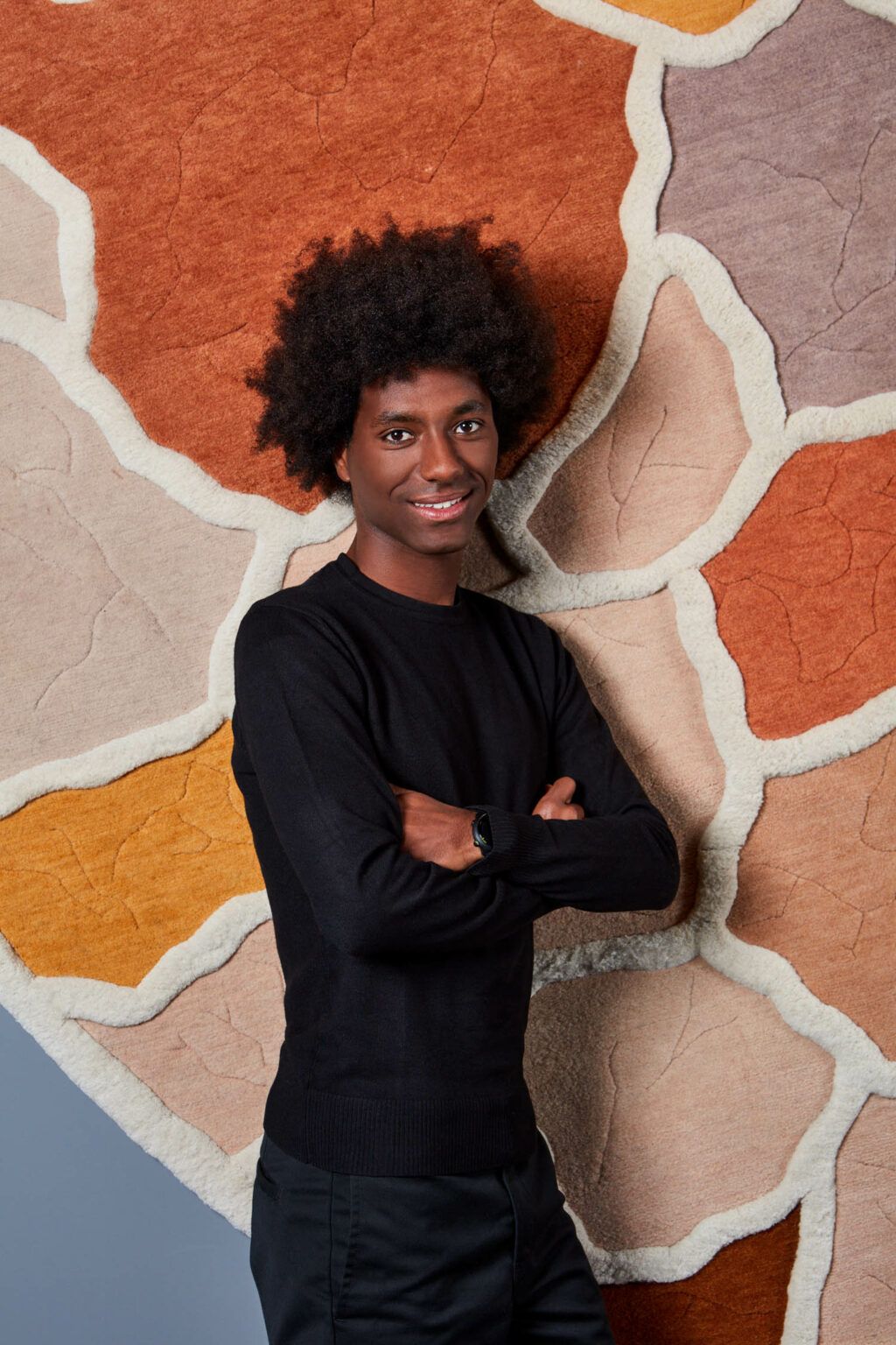 A young man dressed in total black clothing stands in front of an imaginatively designed rug featuring multiple pastel colors.
