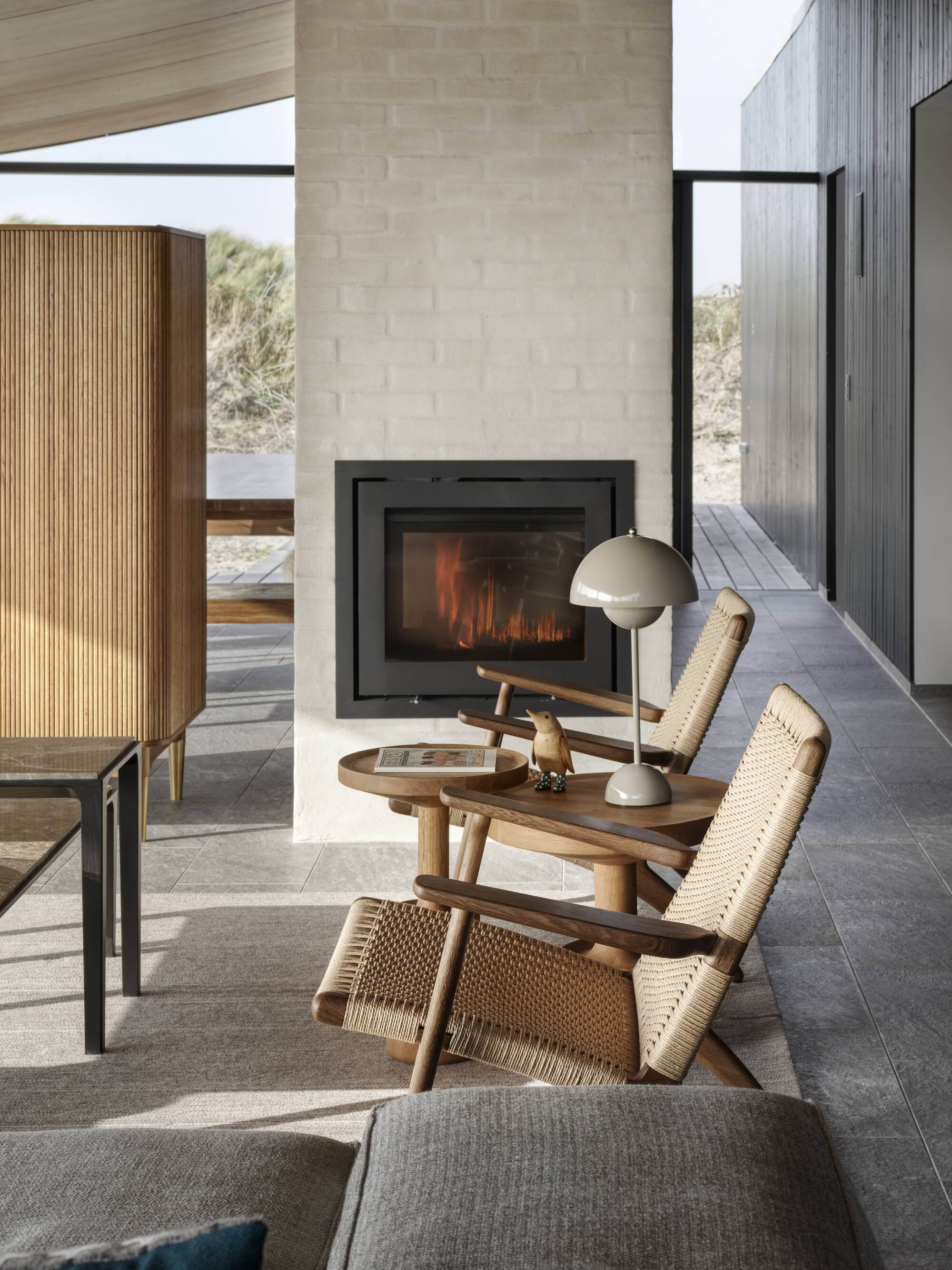 scandi interior with tiled floor, rendered brick freestanding fireplace, two rattan chairs, a gray sofa, a fluted timber freestanding cupboard and a sloped ceiling
