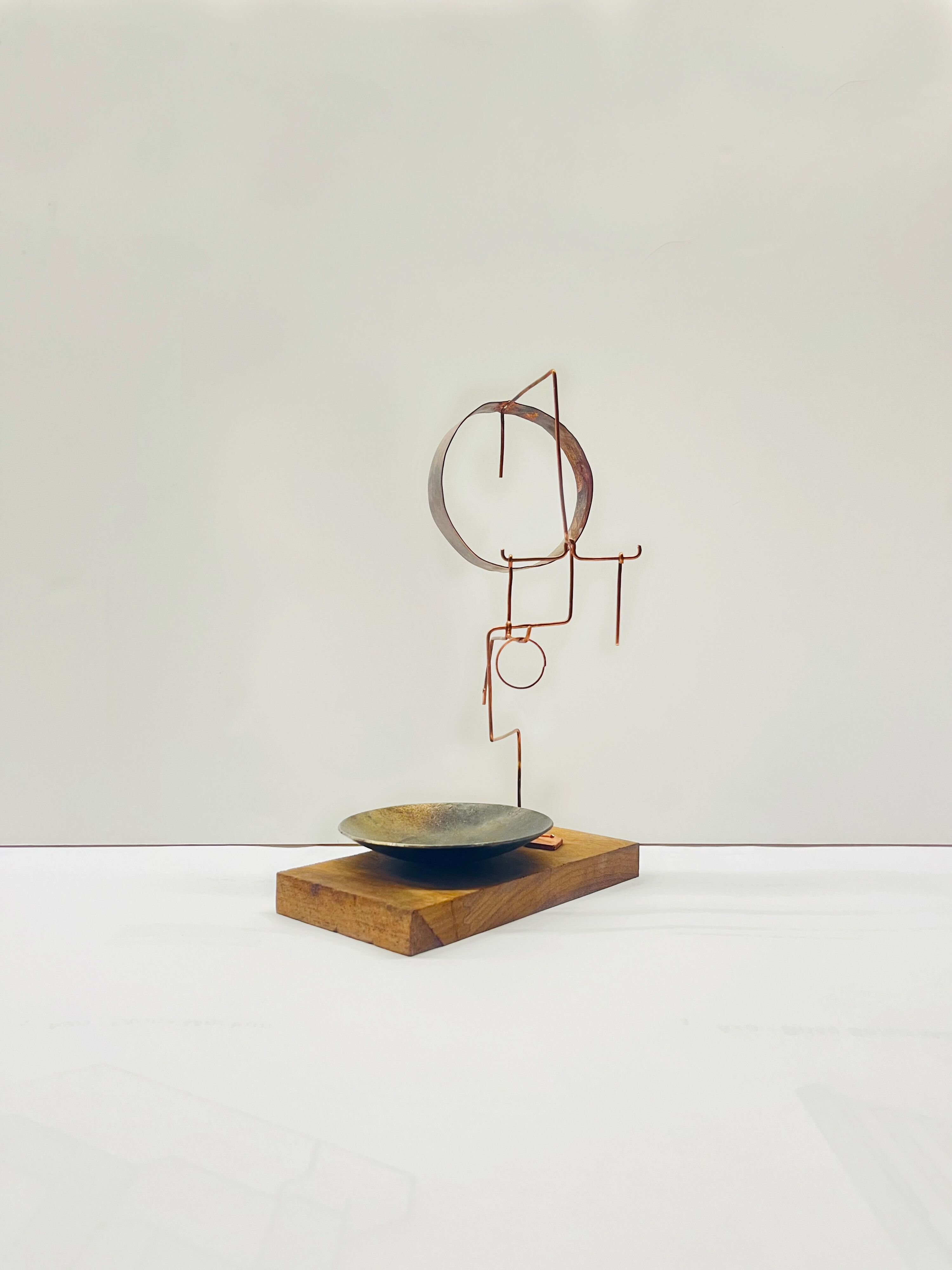 A minimalist birdhouse with a wooden base, a ceramic dish, and an iron-looking, bronze-tinted sculptural totem made of thin geometric shapes sits in a white room.