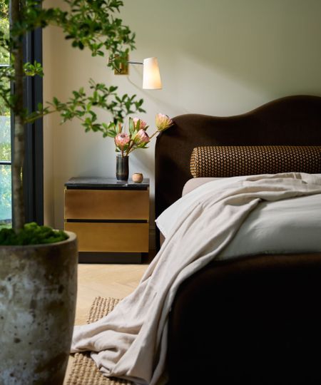 Modern bedroom with curved velvet headboard, marble and metal nightstand with flowers, wall lamp above. Wooden floor, large planter in foreground.
