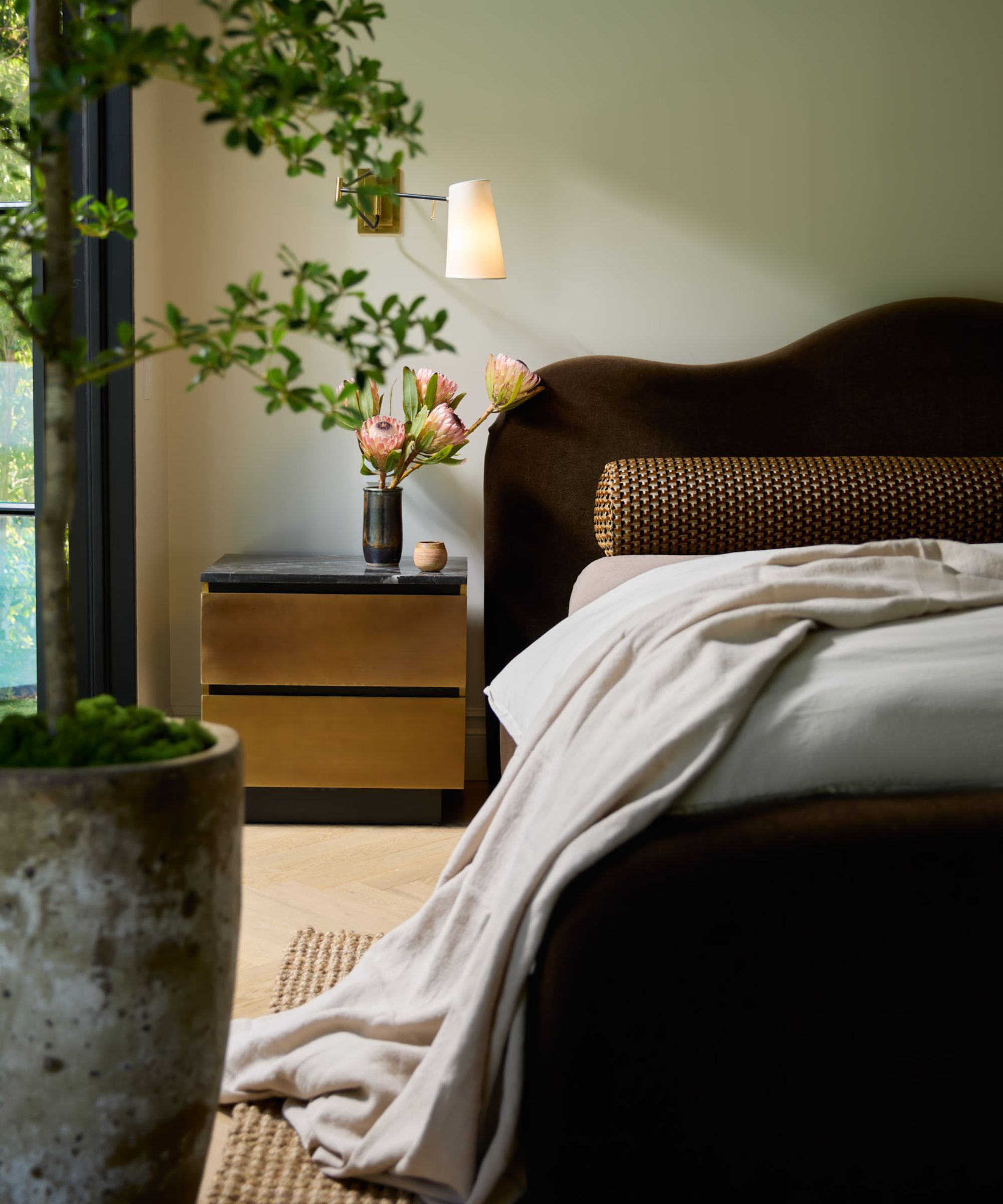 Modern bedroom with curved velvet headboard, marble and metal nightstand with flowers, wall lamp above. Wooden floor, large planter in foreground.