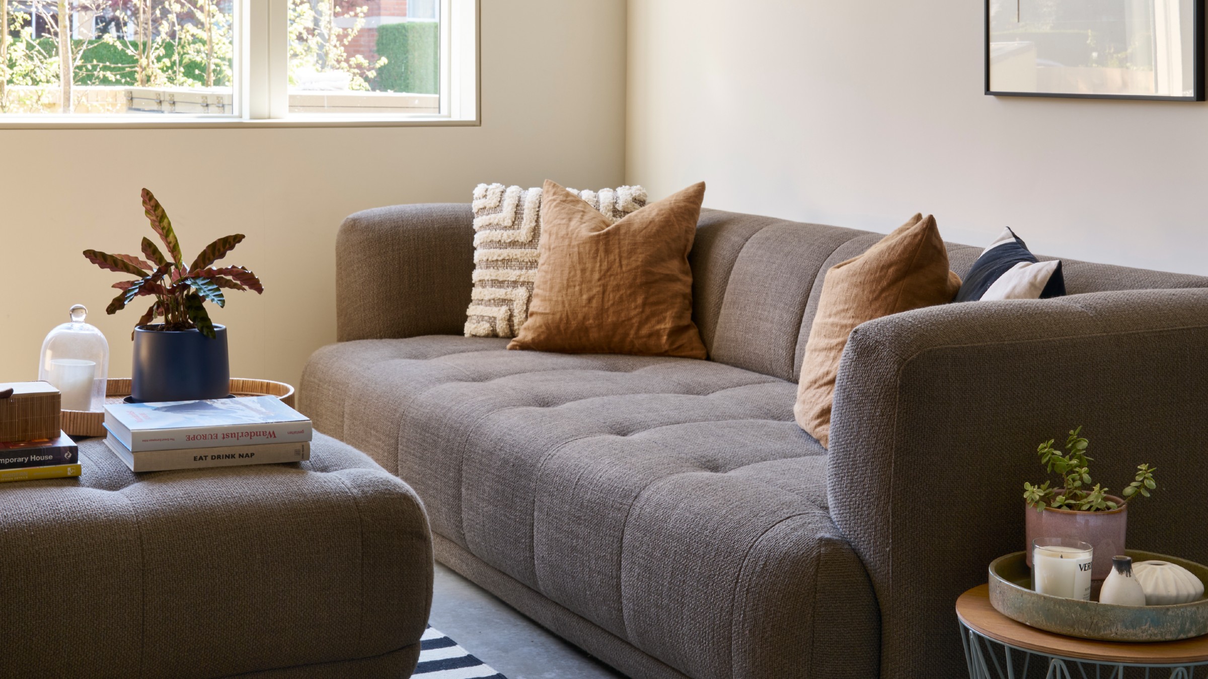 An off-white painted living room with a brown sofa and matching ottoman used as a coffee table