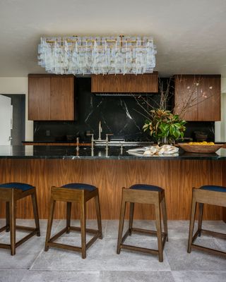 kitchen island with dark wood island cabinets, black backsplash and pale floor tiles four bar stools with black seat cushions and intricate clear glass ceiling light above island