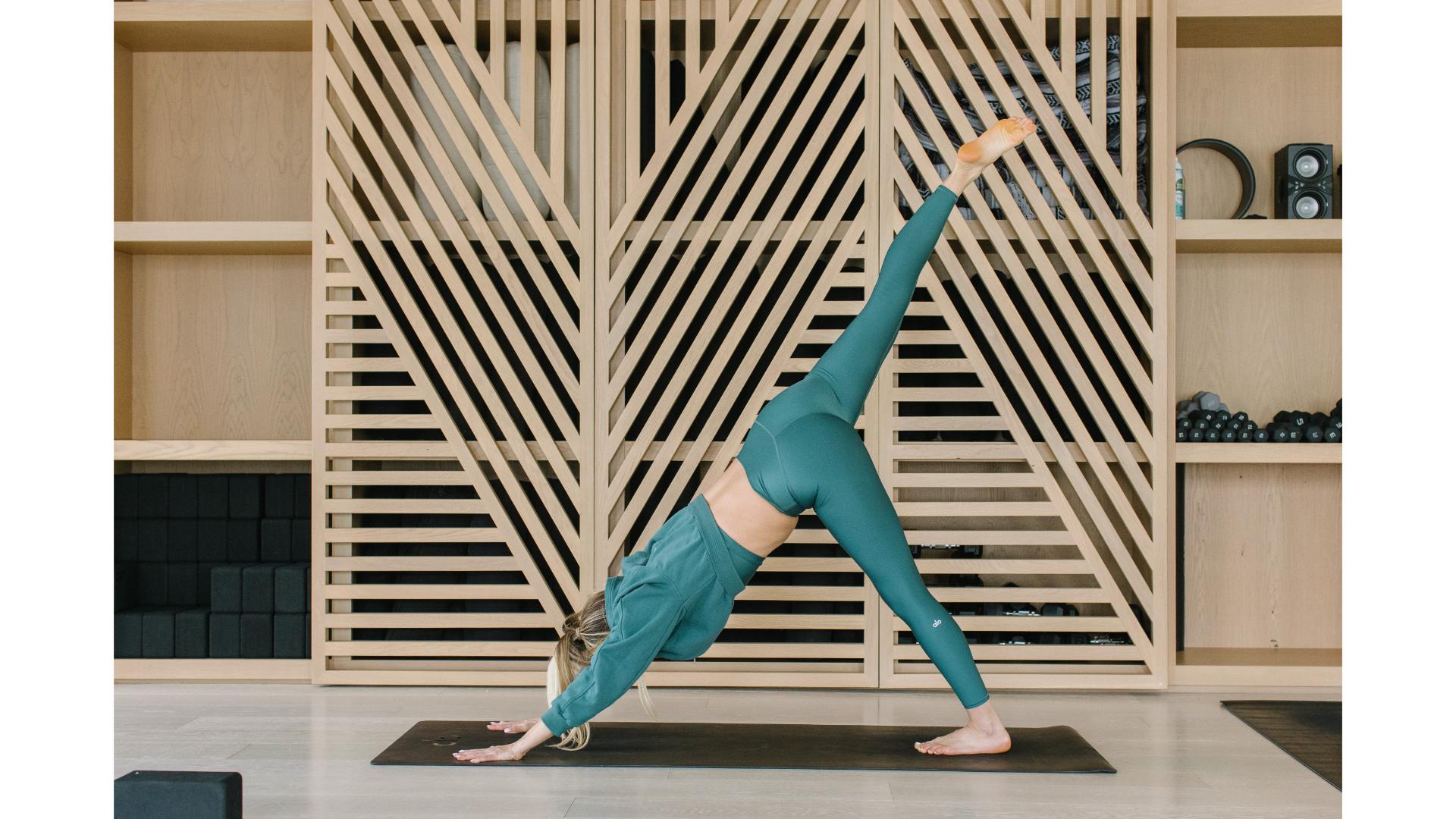 woman dressed in jade green performing a downward facing dog sideways to the camera in with wooden shelving behind her n a studio setting