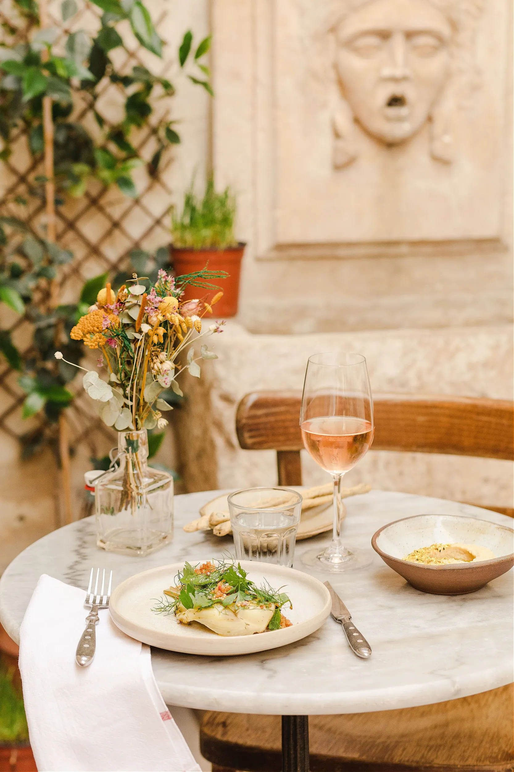 A bucolic food hotspot boasts round marble tables with silver tableware, dry flowers in a transparent glass vase, and a glass of rose wine, all photographed against an ancient stone wall with a human face engraved in it.
