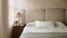 Image of a minimalist bed in a beige, limewash bedroom. There is a wooden side table with a fluted table lamp on it.
