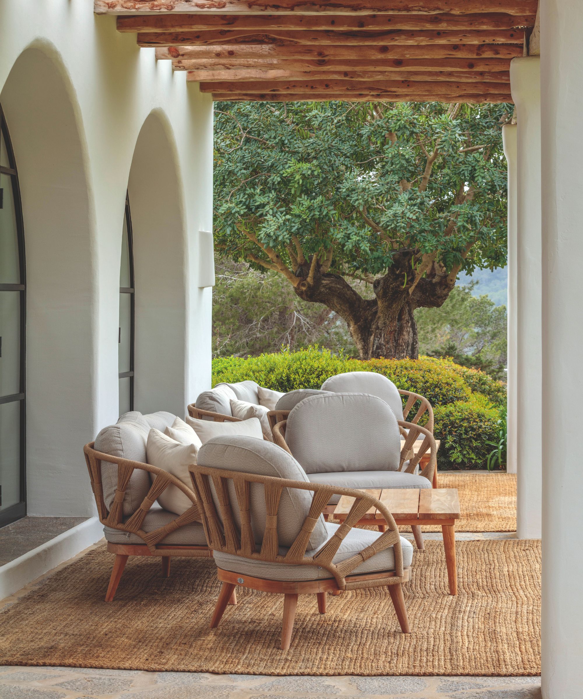 Patio with a beamed ceiling, and white arched walls, a jute rug on the floor, and cane lounge chairs and a wooden table, overlooking a Mediterranean garden.
