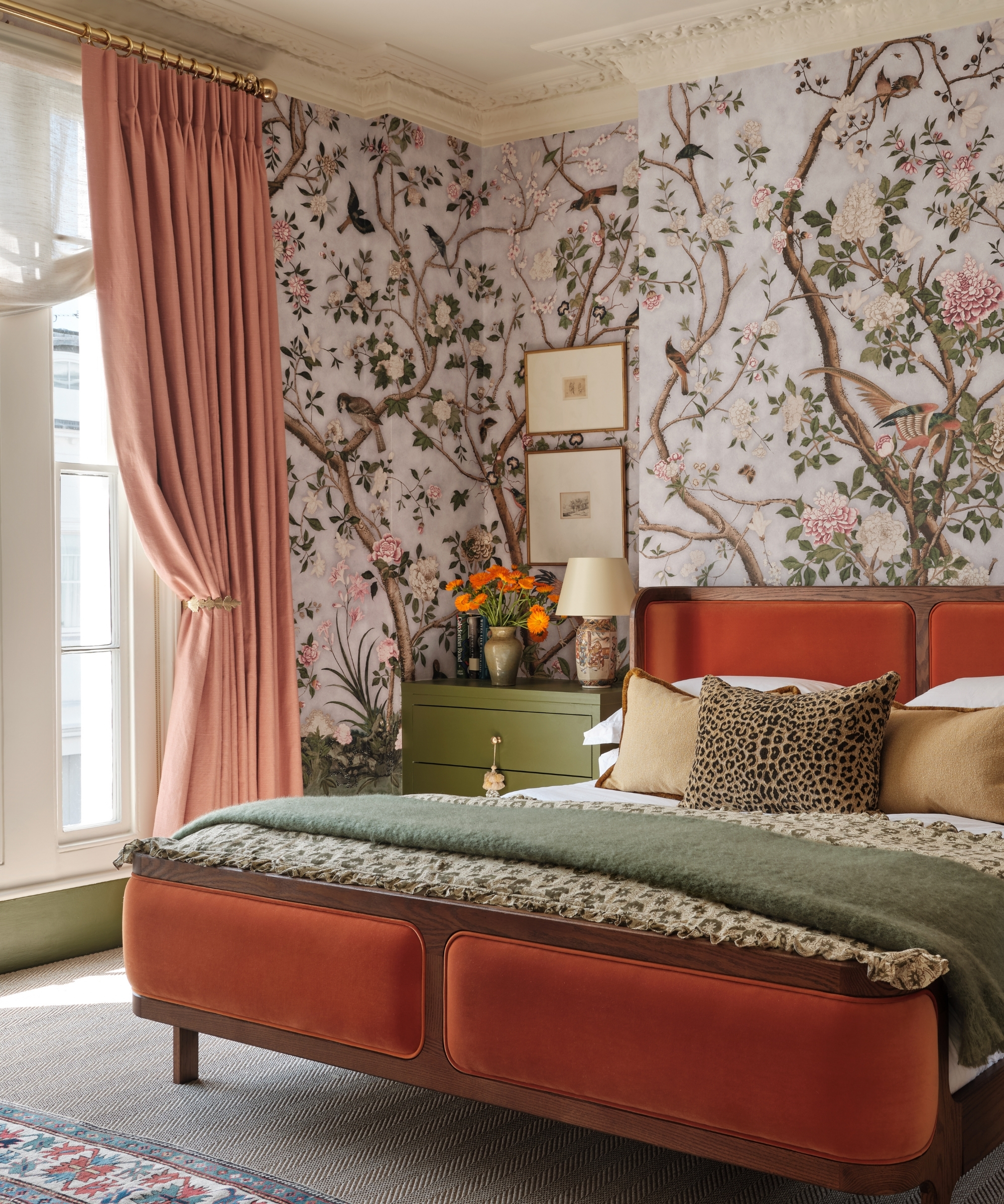 Bedroom with layered sheer blind and pink curtains, floral wallpaper, rust bed and green and patterned textiles.