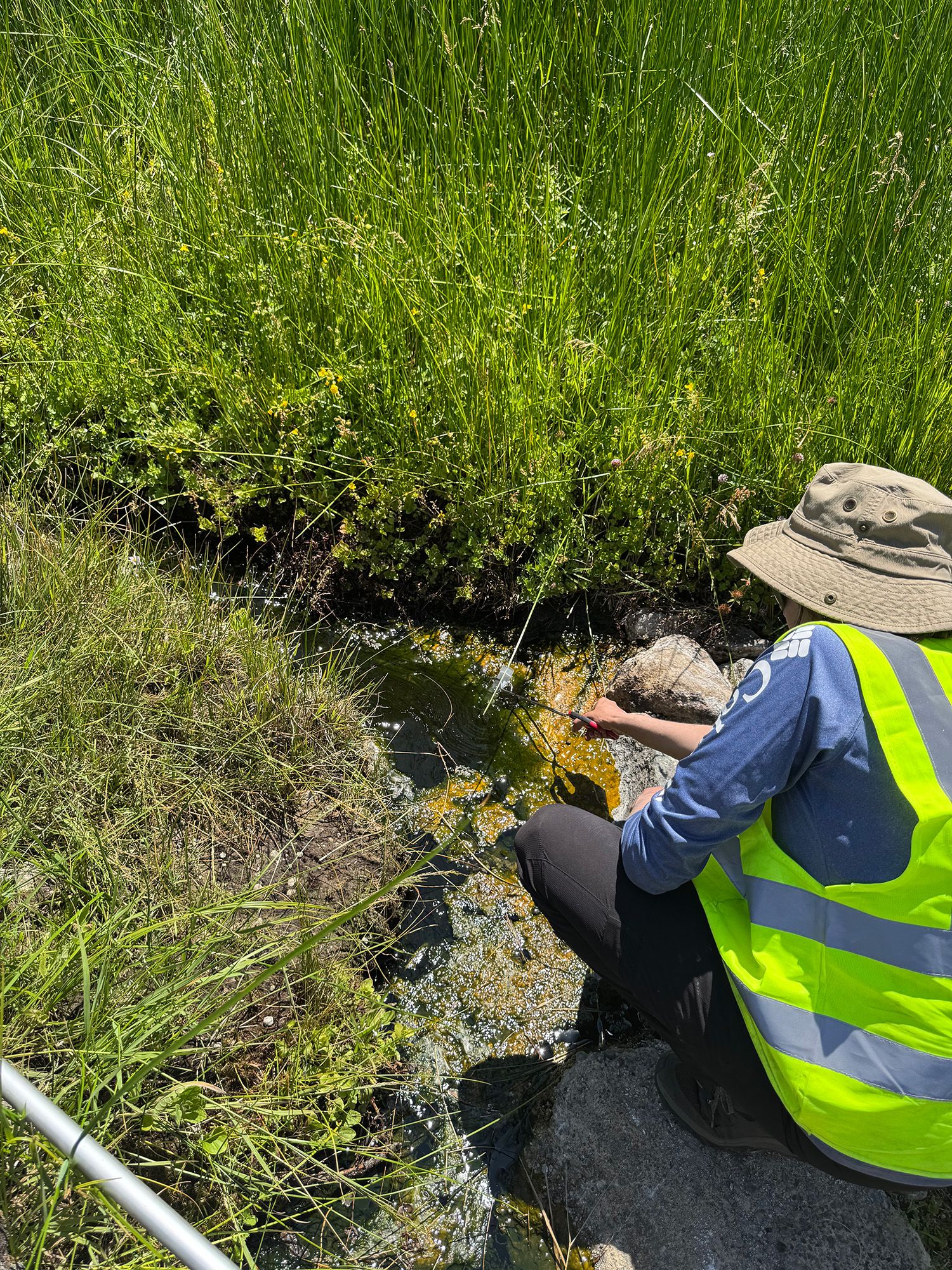 A person wearing a yellow reflective vest and tan bucket hat leans over a small creek.