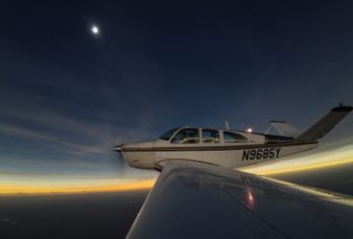total solar eclipse in the top left part of the image photographed from the wing of a airplane which is in the center of the frame.