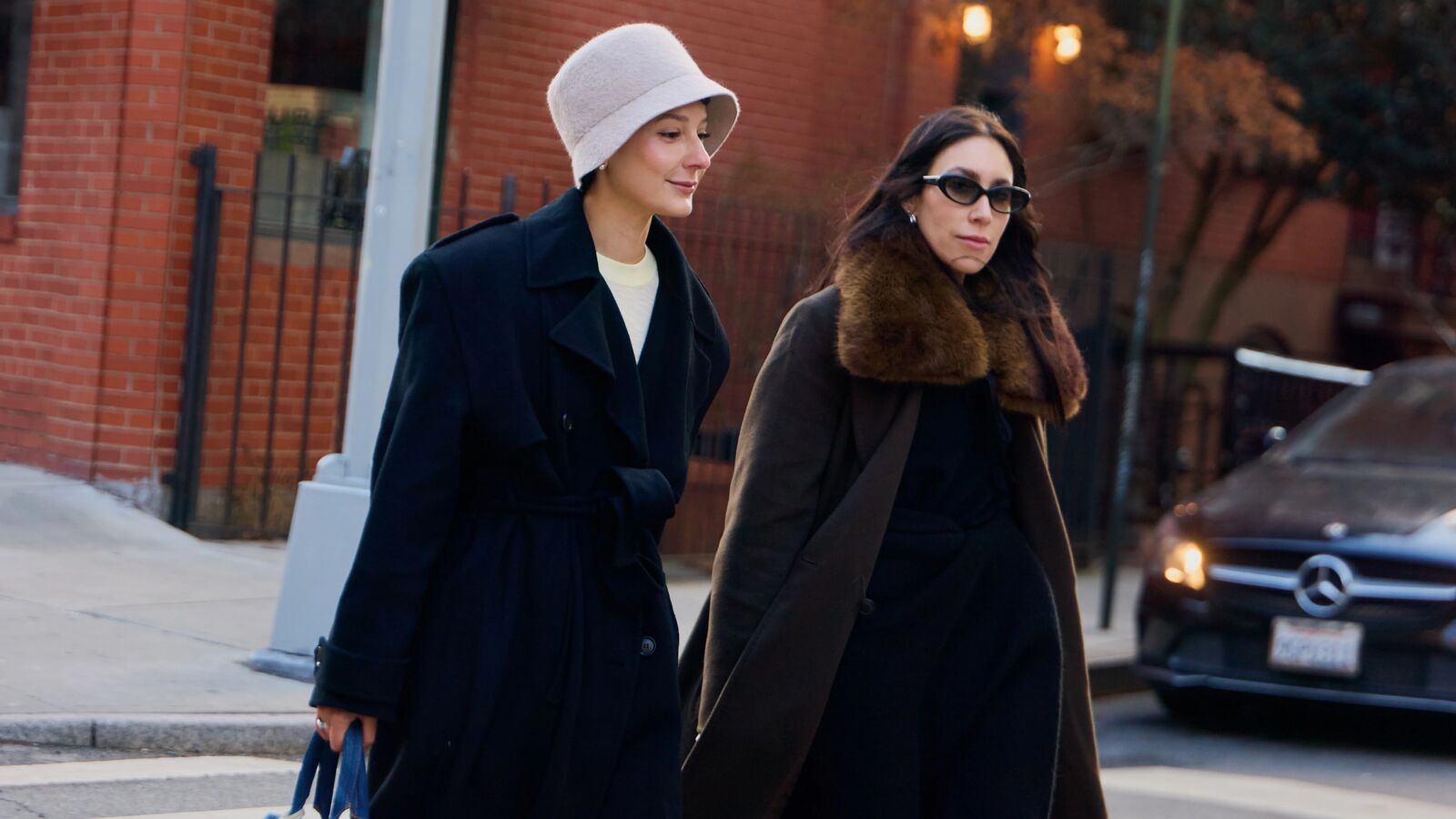Two women are walking on a NYC cross walk. One is wearing a long black coat, bucket hat, black boots, and a blue denim handbag. The other woman is wearing a long black coat with faux fur trim on the collar and tall tan wedged boots. 