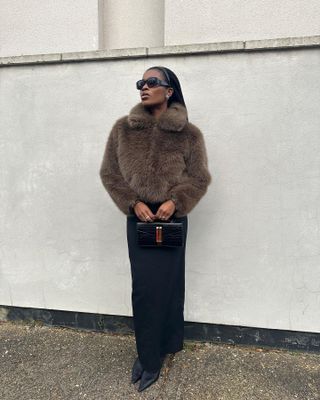 A woman wears a brown short fur coat, black maxi pencil skirt, black pointed toe boots, and a black and gold handbag.