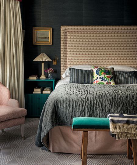 a bedroom with dark walls, a patterned headboard, a gray coverlet on the bed, a soft pink bed skirt, a pink accent chair, a teal bedside table with a lamp and books on it, and a teal bench at the foot of the bed