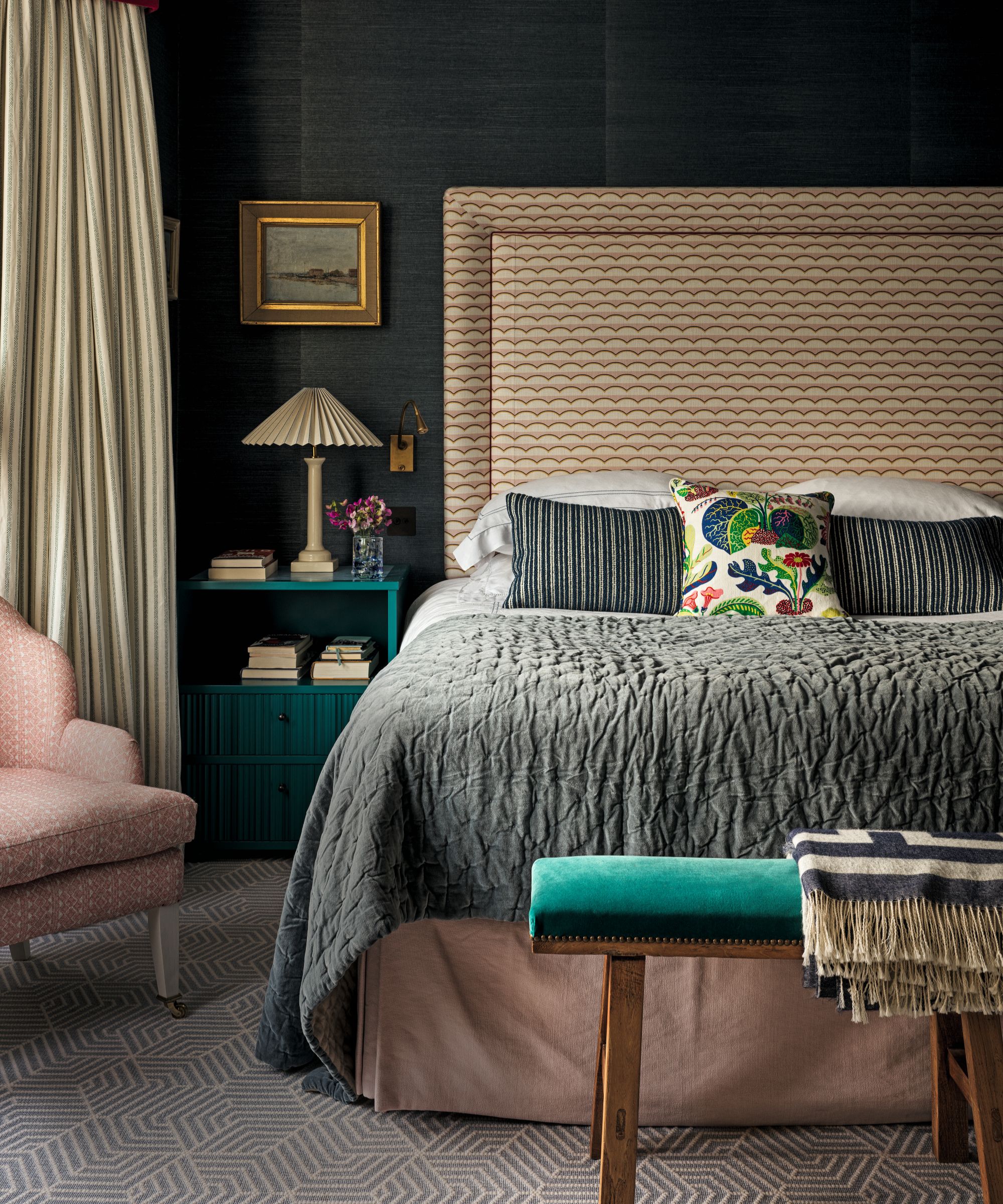 a bedroom with dark walls, a patterned headboard, a gray coverlet on the bed, a soft pink bed skirt, a pink accent chair, a teal bedside table with a lamp and books on it, and a teal bench at the foot of the bed