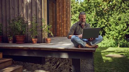 retired man sits on the porch of his tiny home working on his laptop