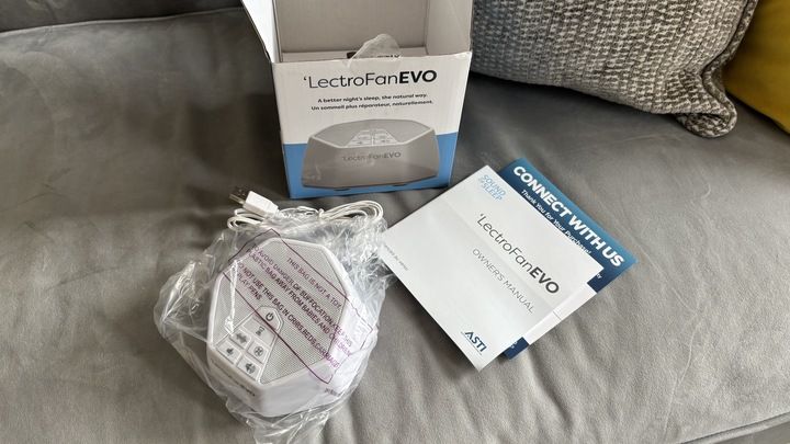 The LectroFan EVO White Noise Machine being unboxed, revealing an instruction booklet, a white electric cord, and the machine wrapped in plastic.