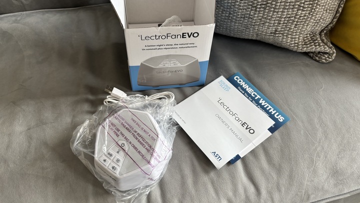 The LectroFan EVO White Noise Machine being unboxed, revealing an instruction booklet, a white electric cord, and the machine wrapped in plastic.