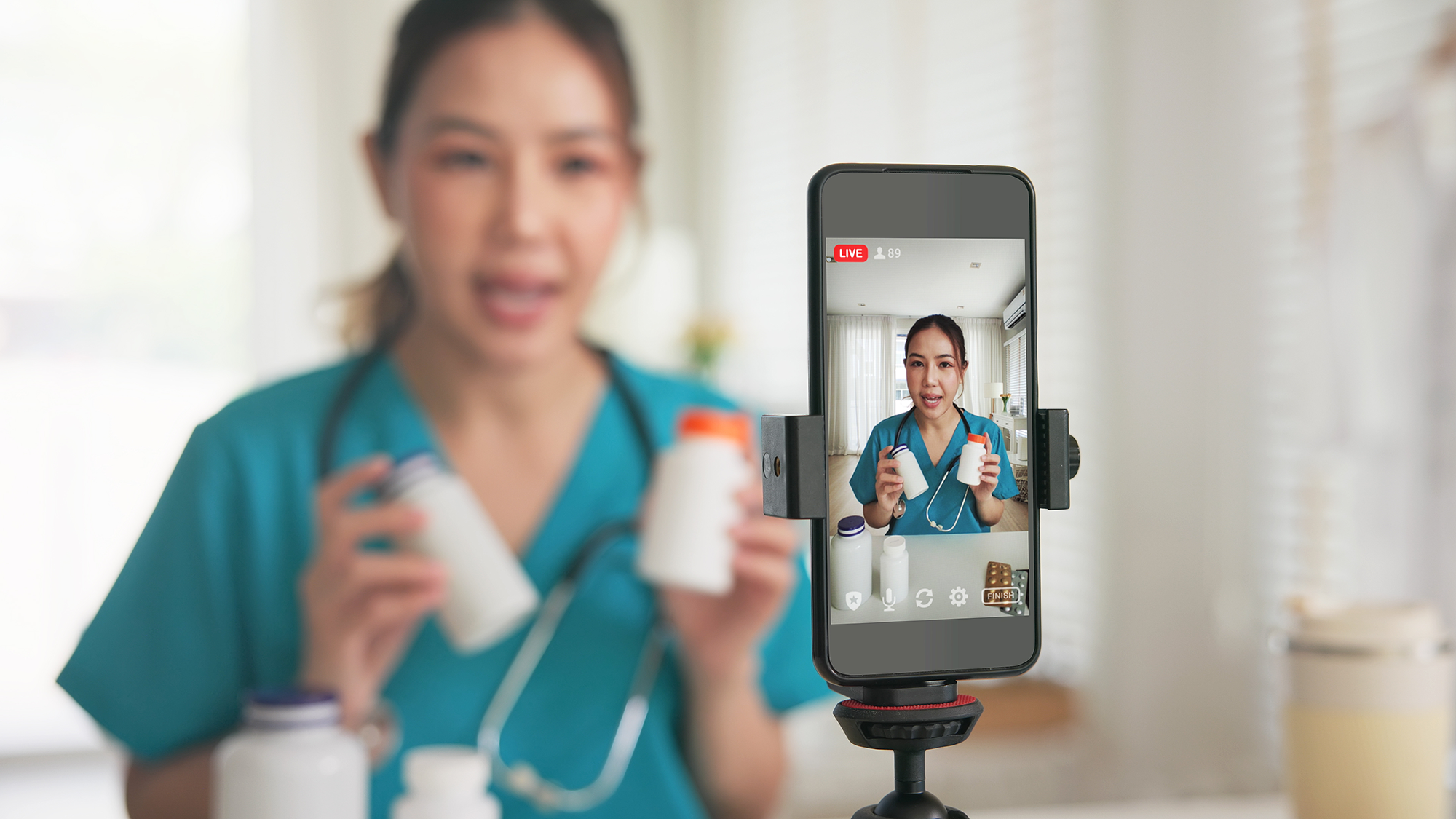 A woman in nurses scrubs with a stethoscope holds two medicine bottles and speaks to a smartphone