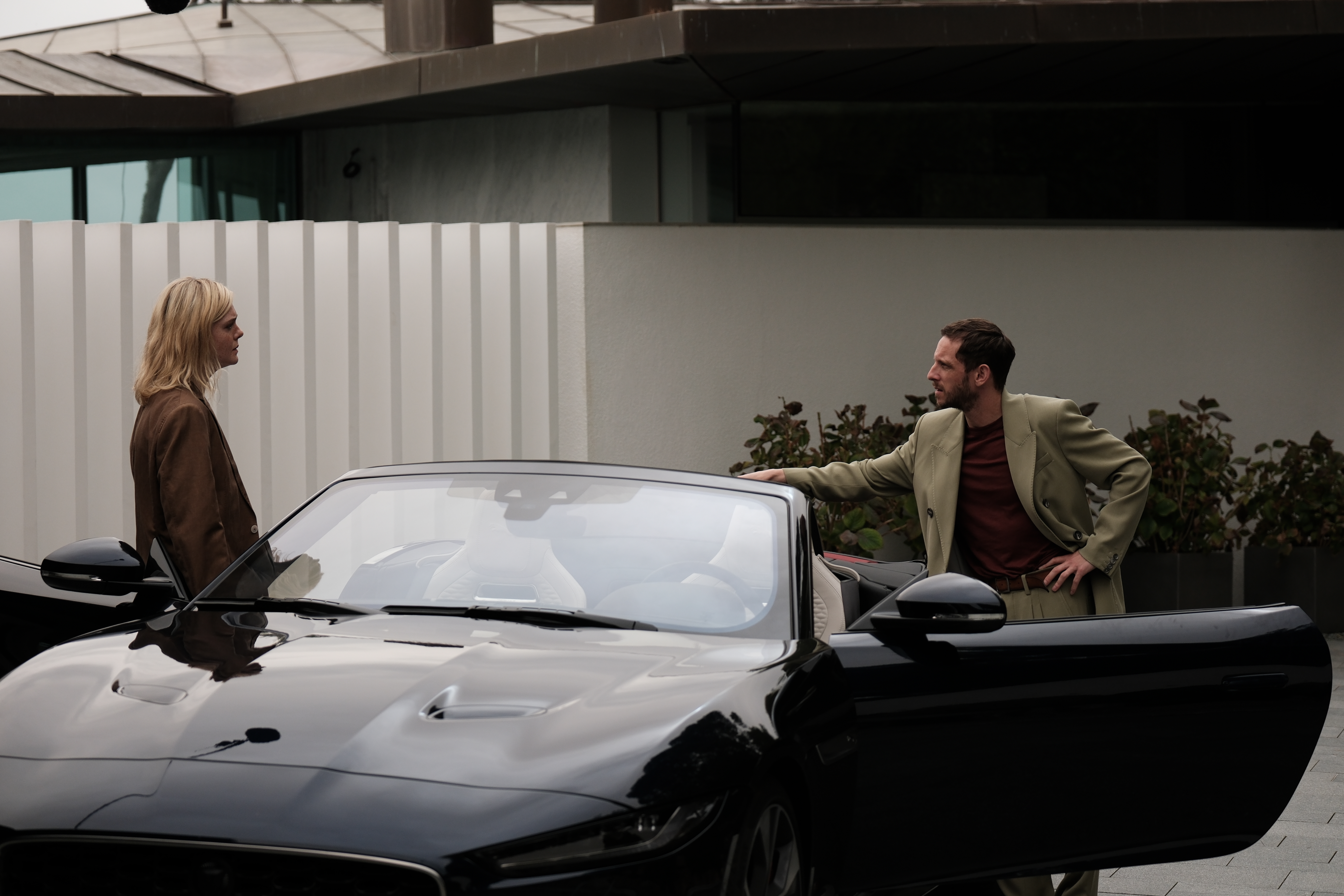 A blond young woman in a brown suit and a man in a pale green one with a brown T-shirt underneath stand next to a sleek black car in front of a gray wall.