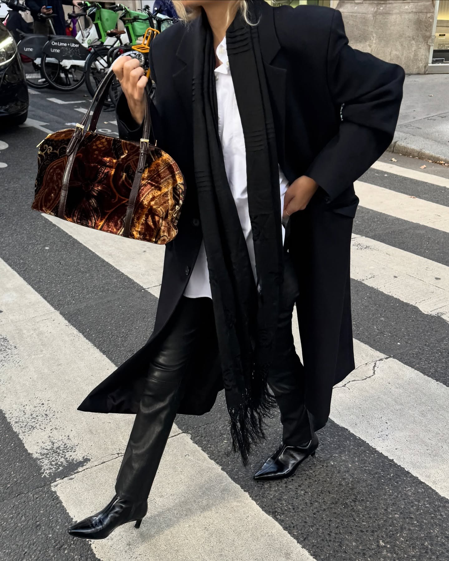 Stephanie wears a long black coat, white button-up, black scarf, black leather pants, and black angkle boots.