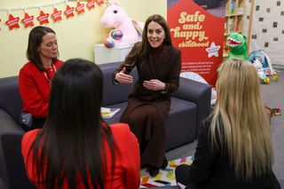 Kate Middleton wearing a long plaid coat sitting on a sofa talking to three women with toys in the background