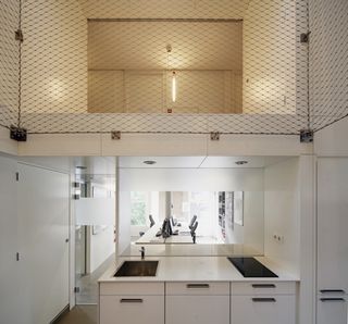Interior view of a cooking area at the Super Cube featuring white walls and white drawers and cupboards with a sink and hob on top. A workspace can also be seen in a separate area along with an upper level that is sectioned off by mesh